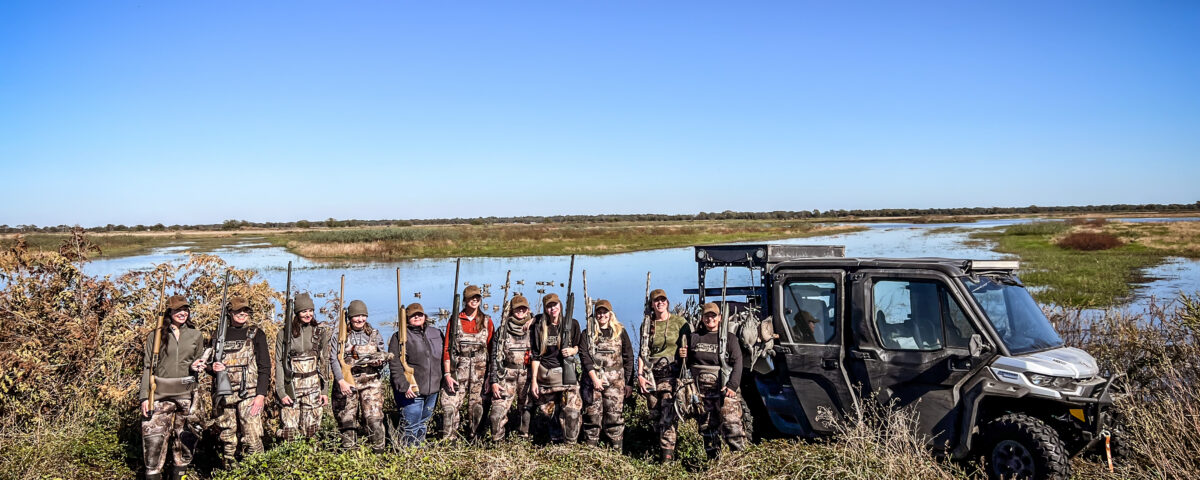 A group of women mentors and their mentees pose in front of a wetland hunting area after a fun day of hunting.