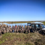 A group of women mentors and their mentees pose in front of a wetland hunting area after a fun day of hunting.