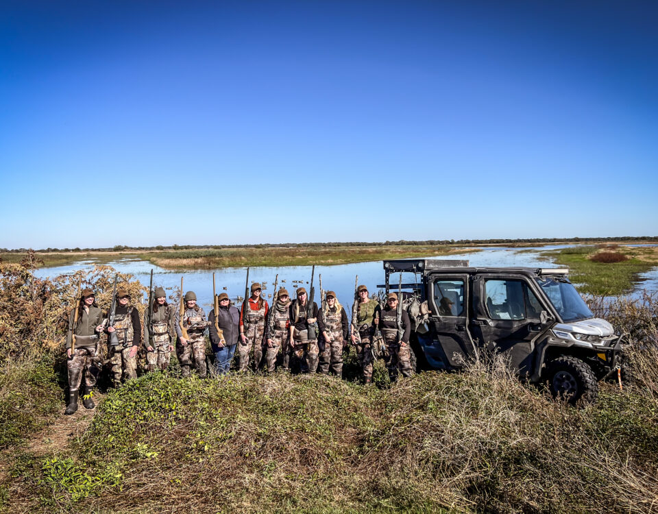 A group of women mentors and their mentees pose in front of a wetland hunting area after a fun day of hunting.